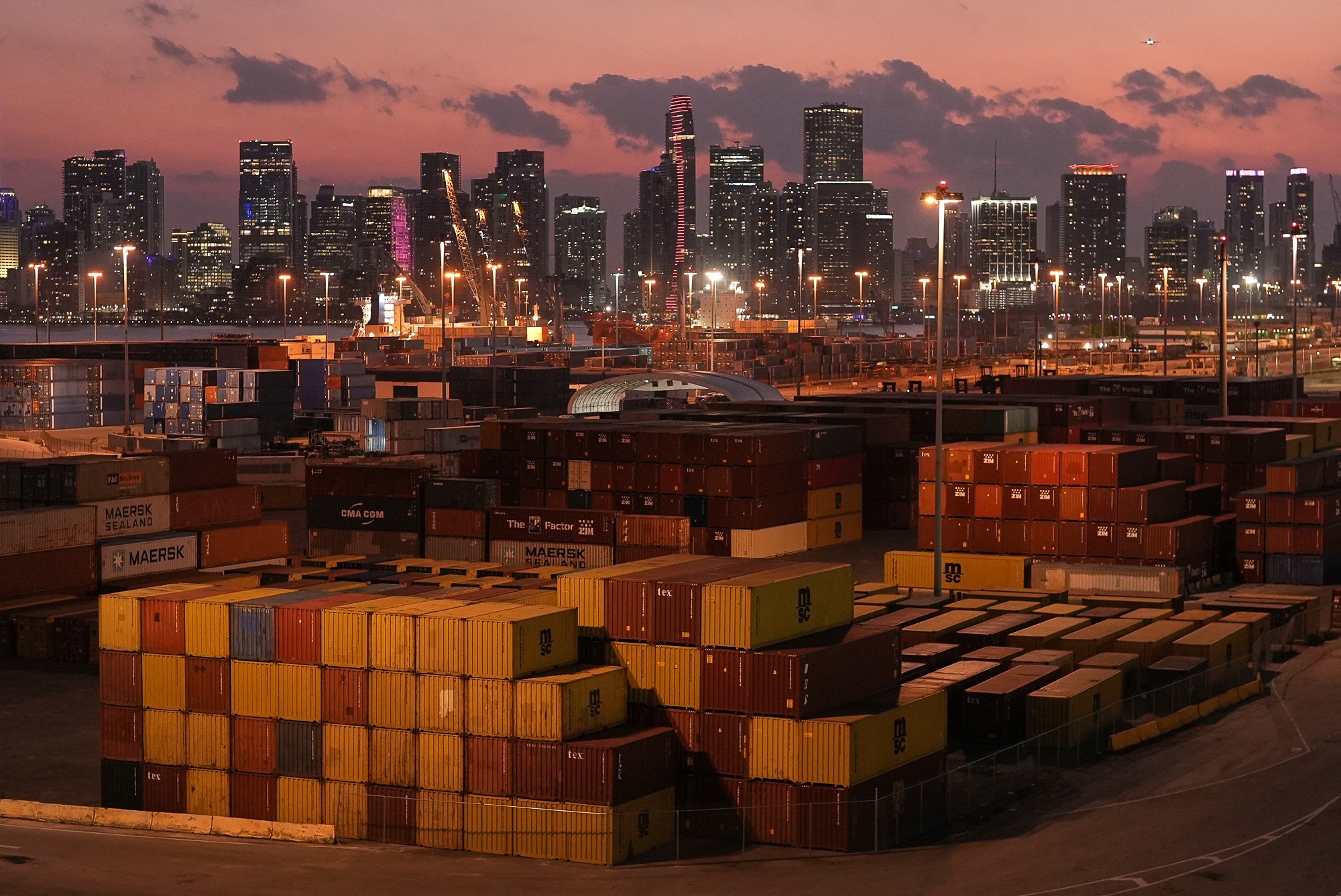 Stacks of shipping containers at a U.S. port at sunset with city skyline in background, symbolizing the impact of tariffs on international trade and consumer prices