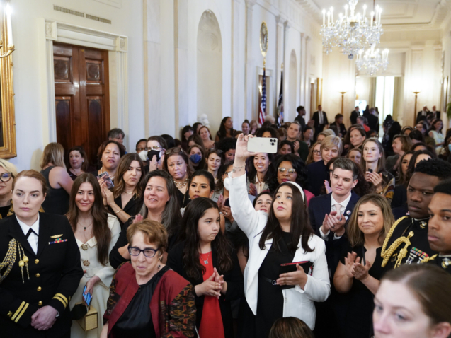 People stand in the Cross Hall of the White House and watch as President Joe Biden speaks during an event to celebrate Equal Pay Day and Women's History Month in the East Room, Tuesday, March 15, 2022, in Washington. (AP Photo/Patrick Semansky)