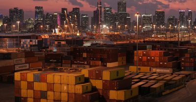 Stacks of shipping containers at a U.S. port at sunset with city skyline in background, symbolizing the impact of tariffs on international trade and consumer prices